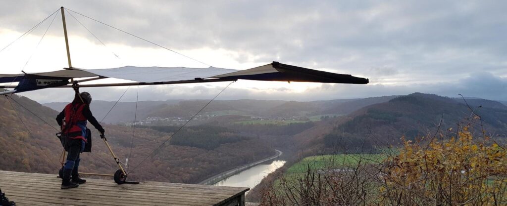 A person standing on a launch platform with a large hang glider. A fall landscape in the distance, sun breaking through the clouds behind the glider, reflection gleaming in the river beside the fields.